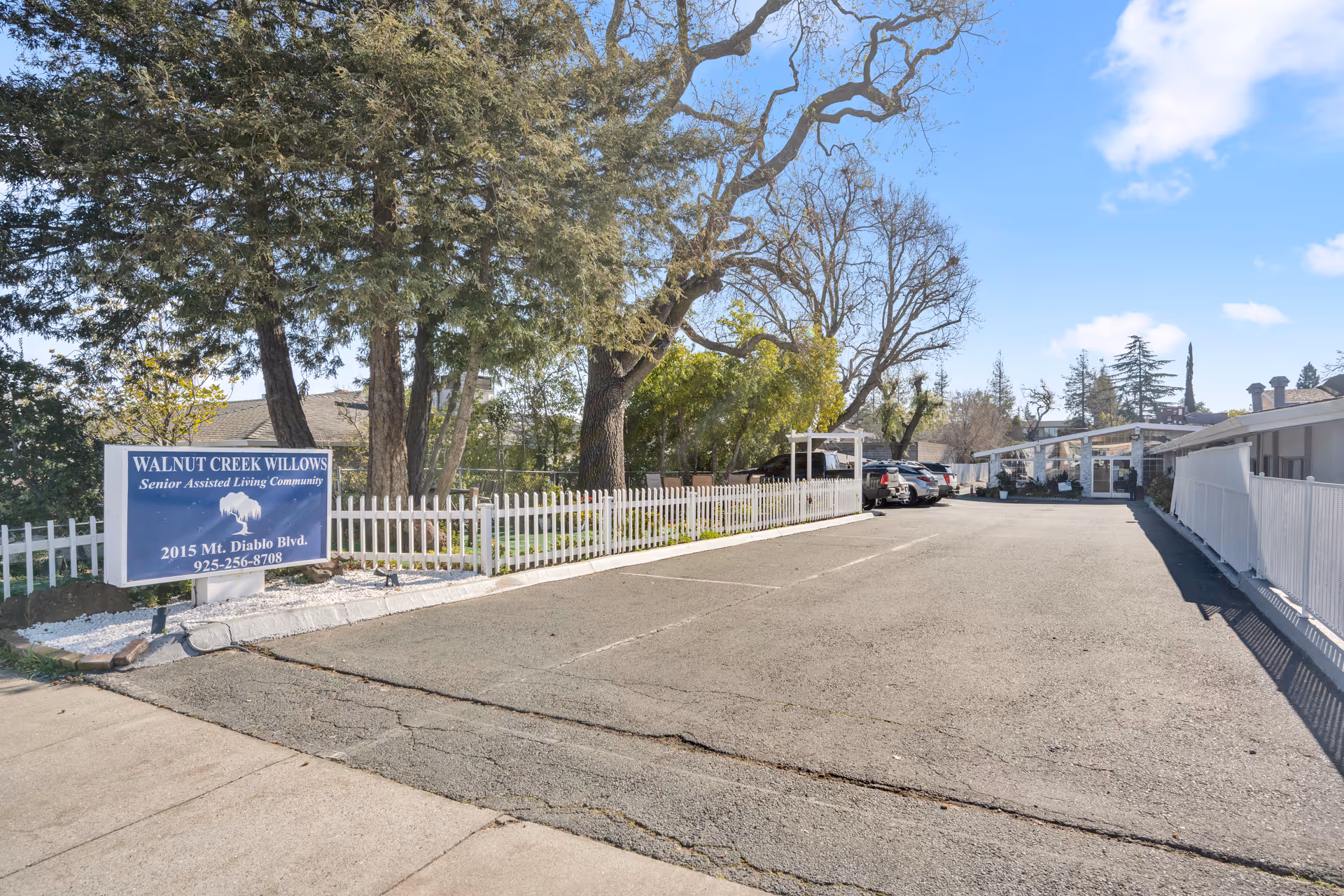 Parking lot entrance with a sign for Walnut Creek Willows senior living next to a white picket fence and large trees.