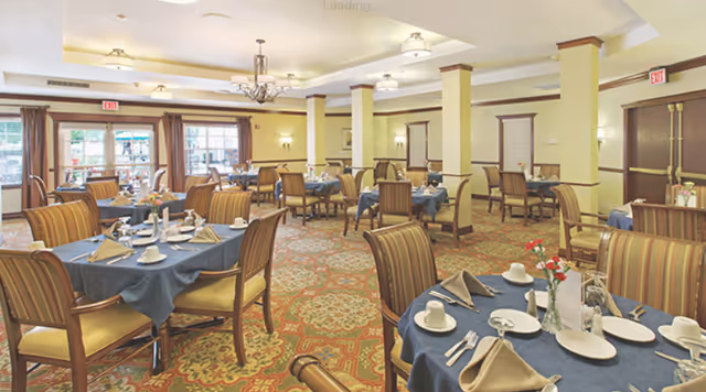 A spacious dining room with multiple tables covered in blue tablecloths and set with white plates, cups, silverware, and beige napkins. The room features patterned carpet, yellow walls with white trim, several columns, and large windows with brown curtains allowing natural light to enter.