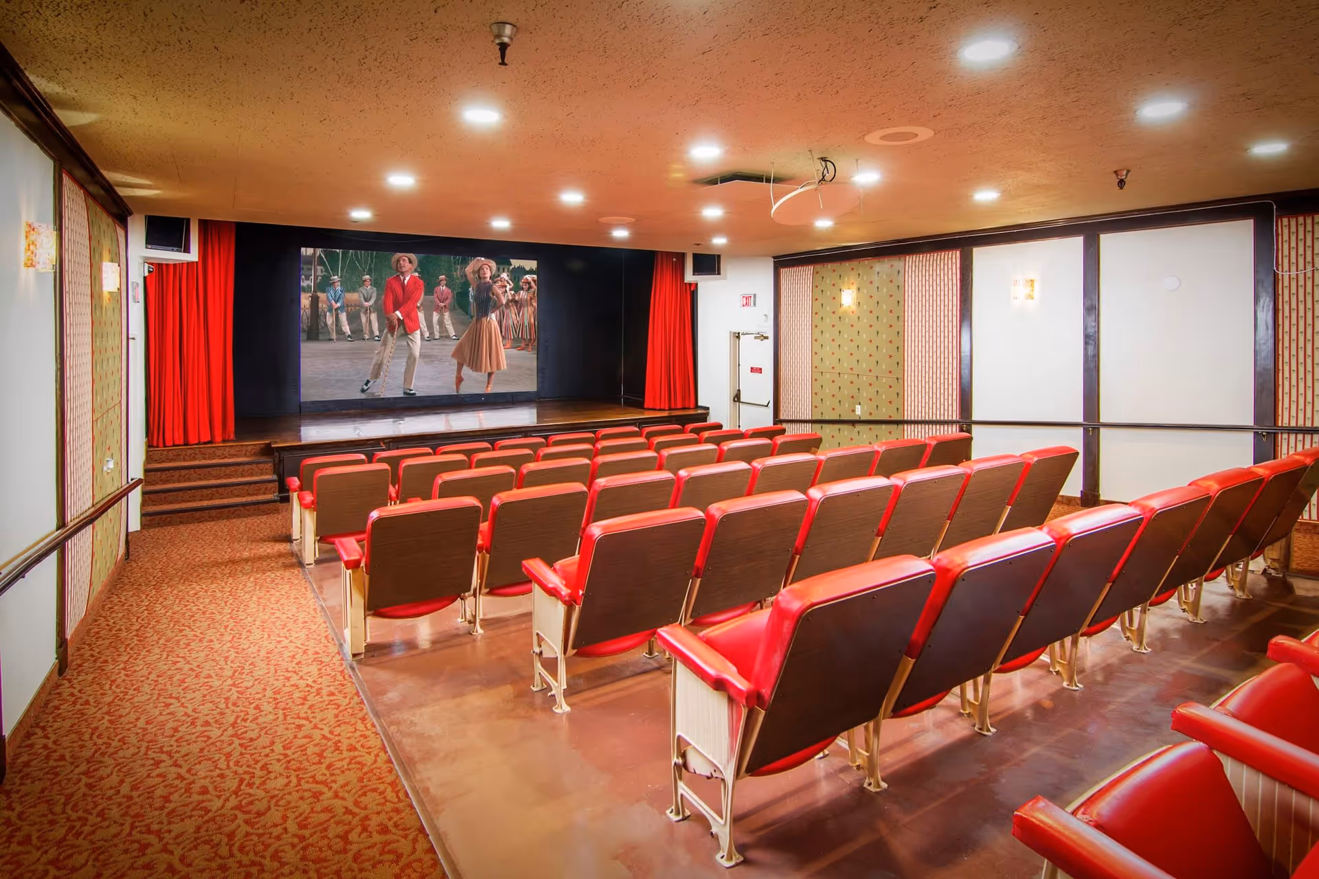 Interior view of a small theater room with rows of red and brown cushioned seats facing a stage with a large screen showing a scene from a musical performance. The room has patterned carpet, red curtains on the stage, and soft ceiling lights.