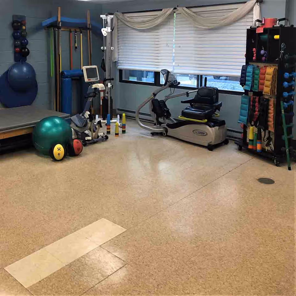 A rehabilitation exercise room with various fitness and therapy equipment including an exercise bike, resistance bands, therapy balls, and a padded therapy table. The room has a large window with blinds and a beige tiled floor.