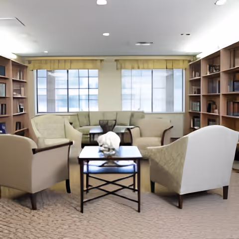 A cozy sitting area with beige armchairs and a matching sofa arranged around a wooden coffee table. The room has large windows with yellow valances, built-in wooden bookshelves filled with books, and a carpeted floor.