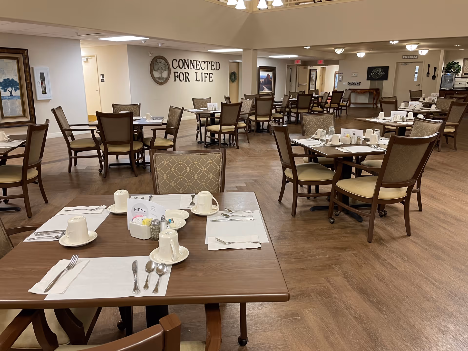A dining room in a senior living facility with multiple tables and chairs arranged neatly. Each table is set with white placemats, cups, saucers, utensils, and a menu holder. The room has wooden flooring and neutral-colored walls. On one wall, there is a decorative clock and the phrase 'CONNECTED FOR LIFE' displayed. The lighting is warm with ceiling fixtures providing illumination.