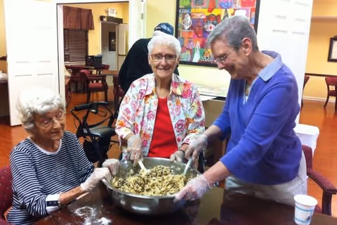 Three elderly women wearing gloves are gathered around a table mixing ingredients in a large metal bowl inside a senior living community. They are smiling and appear to be enjoying the activity. The room has warm lighting and colorful artwork on the wall in the background.