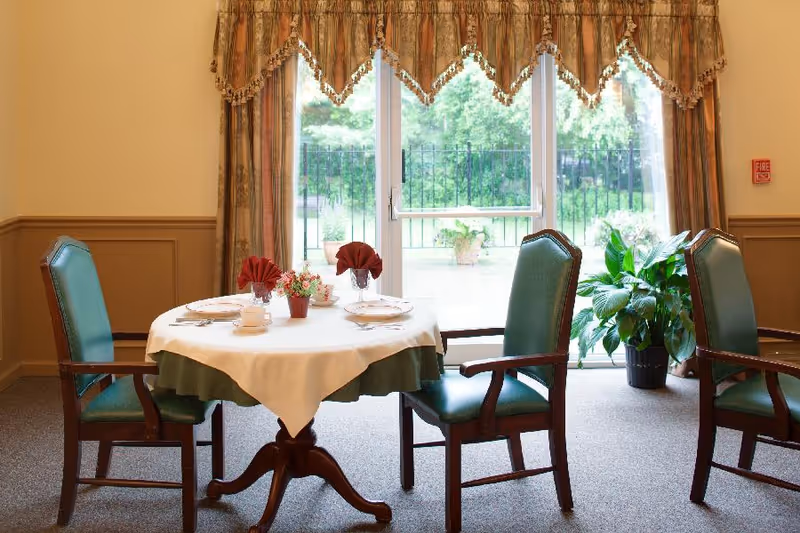 A dining area with a round table covered with a white tablecloth and green underlay, set for two with plates, cups, and folded red napkins. Four green upholstered wooden chairs surround the table. Behind the table is a large window with patterned curtains, showing a view of a garden outside. A potted plant is placed near the window on the right side.
