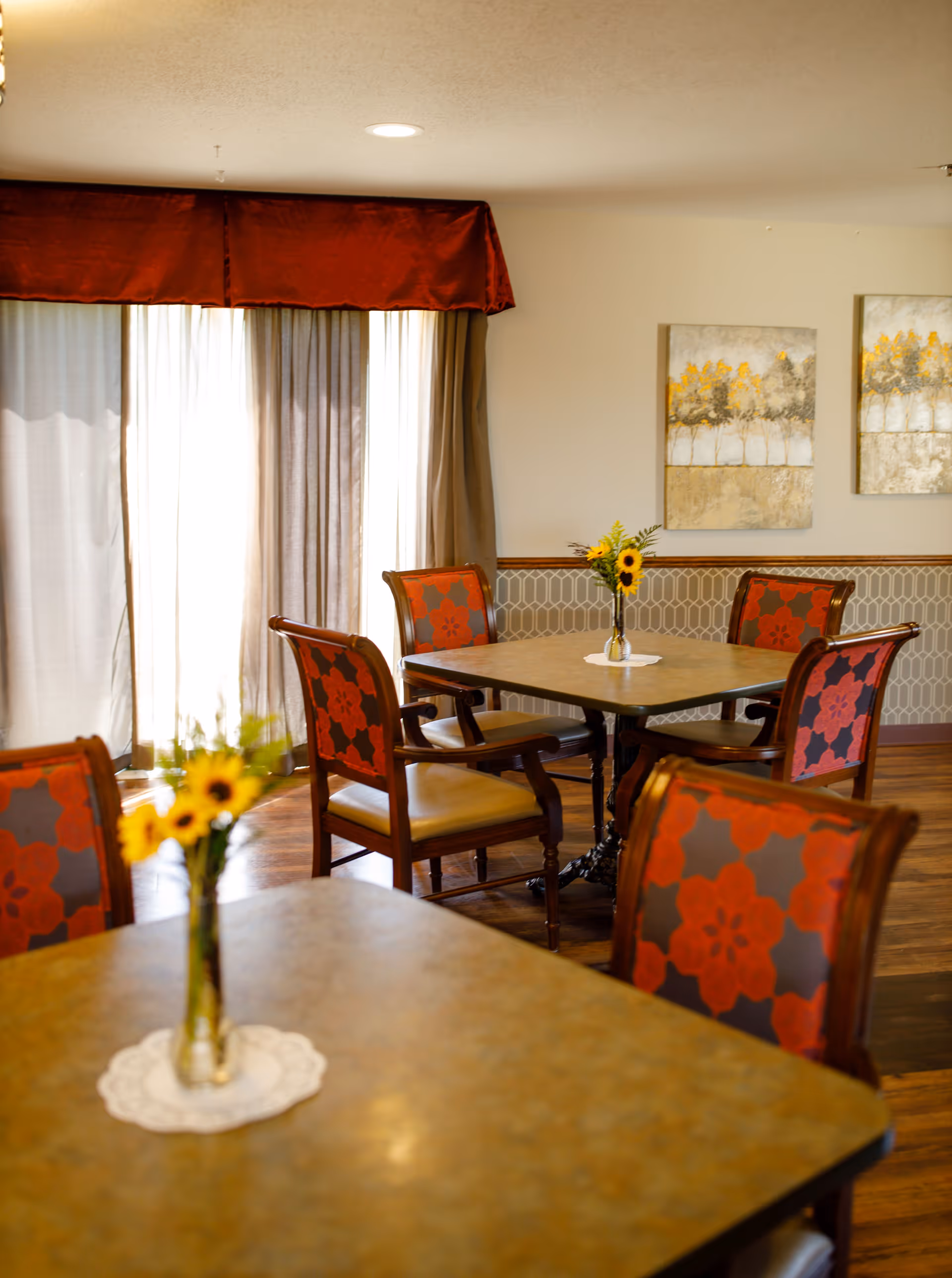 A dining area with square tables and wooden chairs featuring red and gray floral upholstery. Each table has a small vase with sunflowers. The room has large windows covered with sheer curtains and a red valance. Two paintings with abstract tree designs hang on the wall.