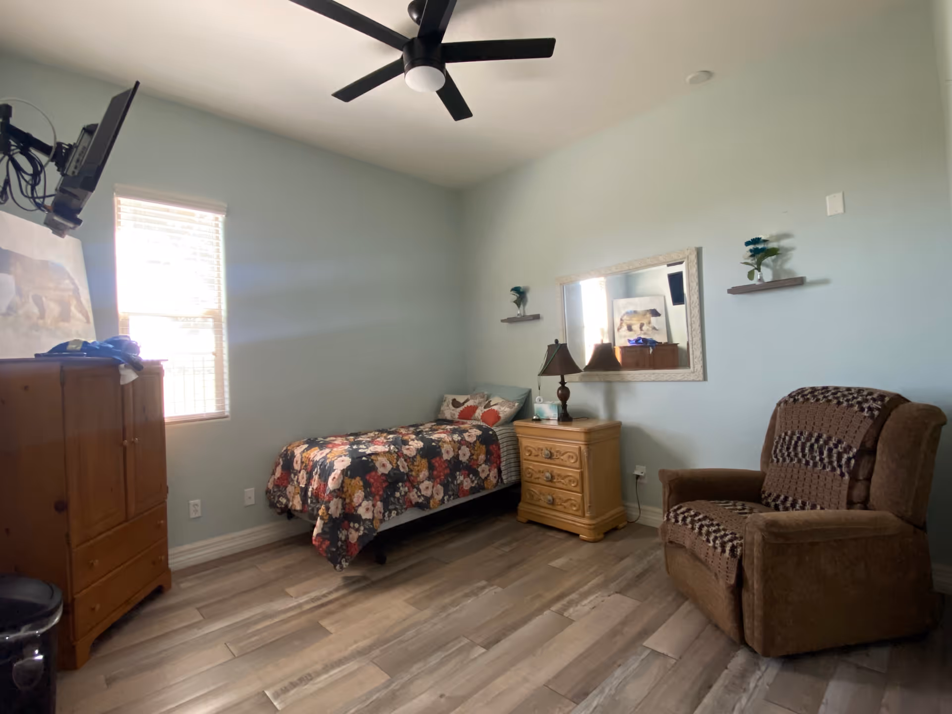 A tidy bedroom with a single bed, wooden dresser, armchair, mirror, and ceiling fan.