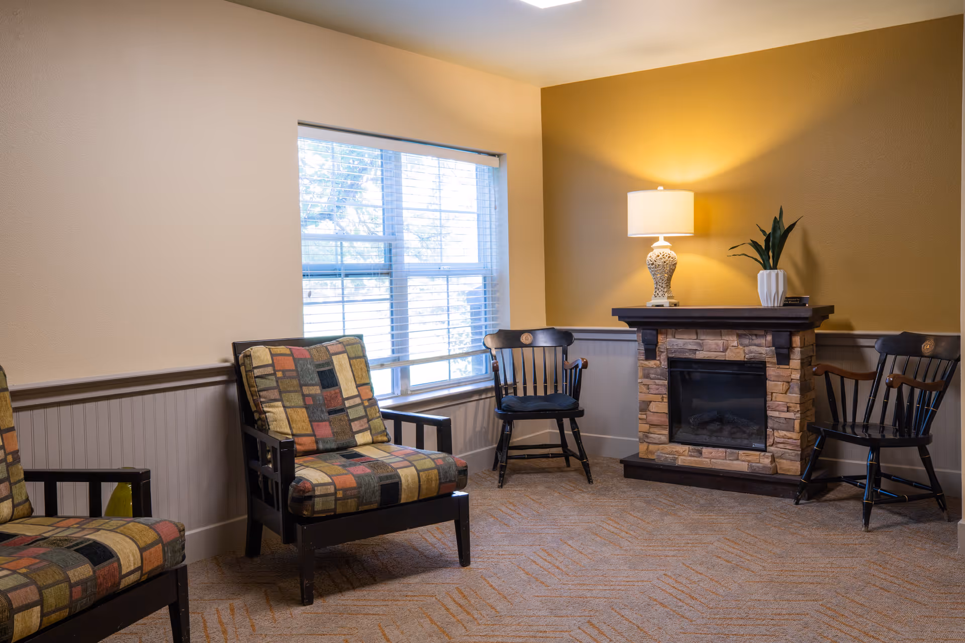 A cozy sitting room with patterned armchairs, a stone fireplace topped by a lamp and plant, and a window letting in daylight.