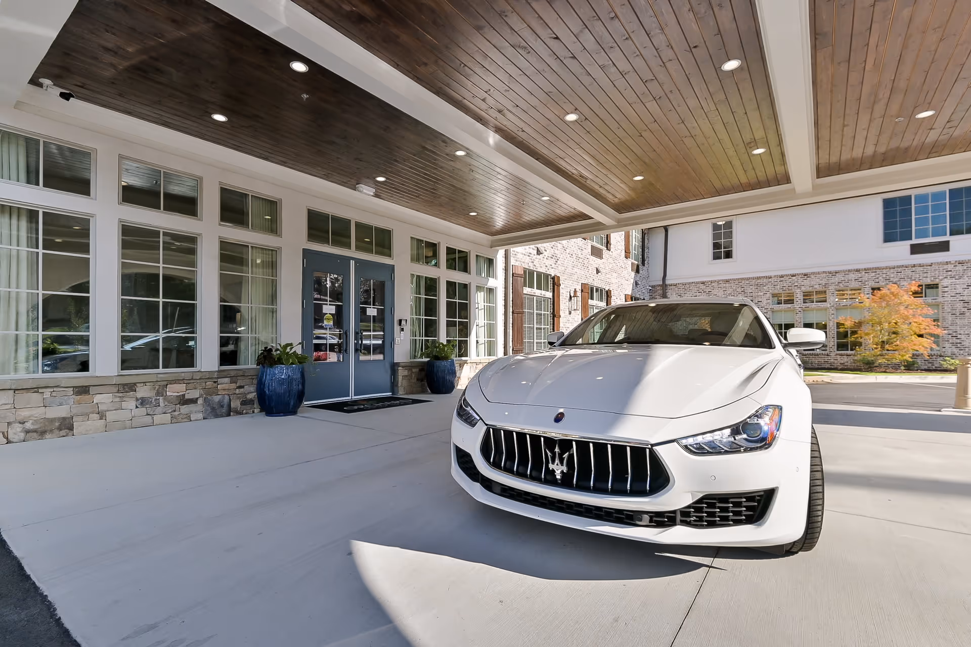 Front entrance of Orchard at Brookhaven facility with a covered driveway and a white luxury car parked in front. The building features large windows, stone accents, and a wooden ceiling with recessed lighting.