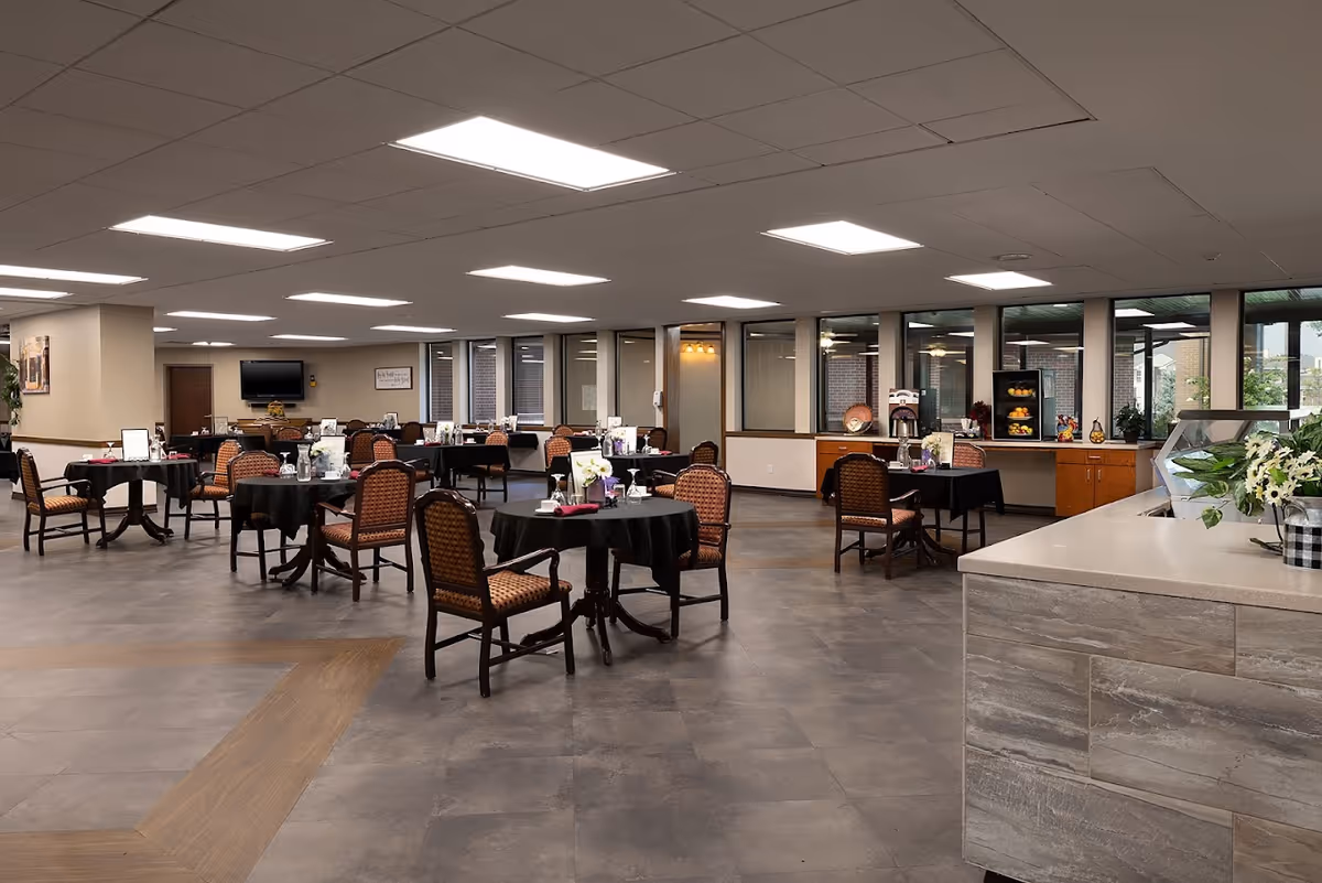 Spacious dining room with multiple round tables covered in black tablecloths and cushioned chairs, set within a senior living facility.