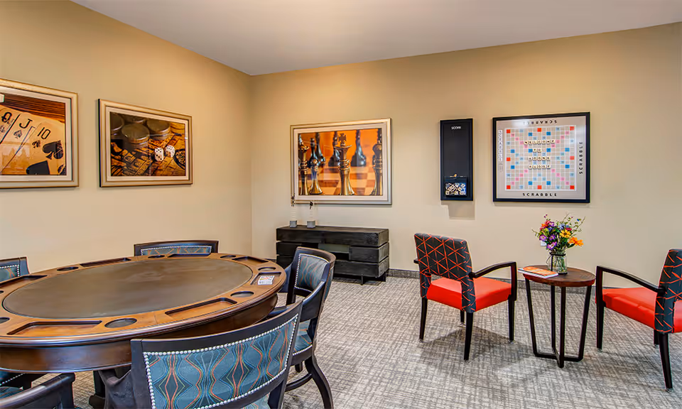 A game room with a round poker table surrounded by chairs on the left and two red and black patterned chairs with a small round table holding a vase of flowers on the right. The walls are decorated with framed pictures of playing cards, chess pieces, and a Scrabble board.