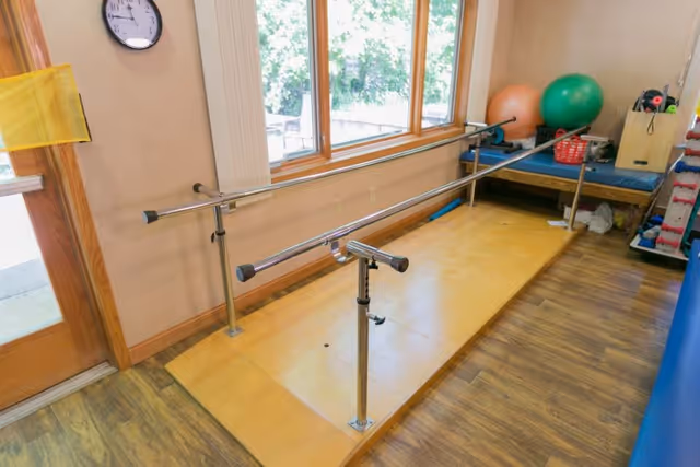 Physical therapy room with parallel bars for walking exercises, a clock on the wall, large windows letting in natural light, exercise balls, and various therapy equipment in the corner.
