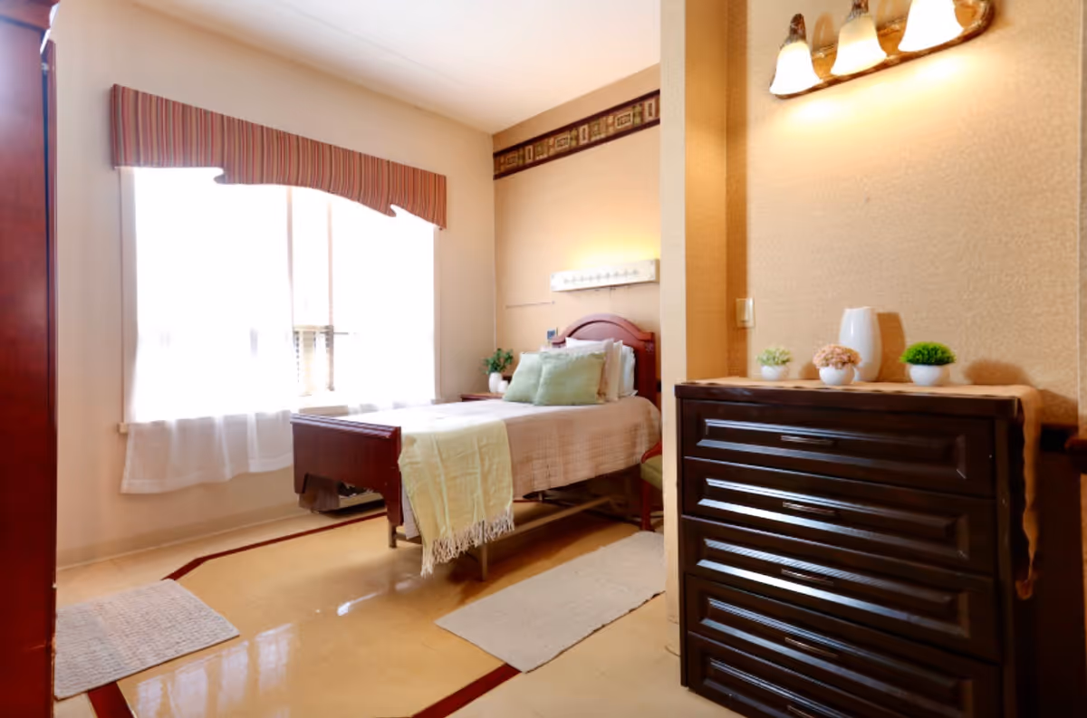 A bright and cozy bedroom in a senior living facility with a single bed covered in light-colored bedding and green pillows. The room has a large window with sheer white curtains and a striped valance, a dark wooden dresser with small potted plants and a white vase on top, and warm lighting fixtures on the wall.