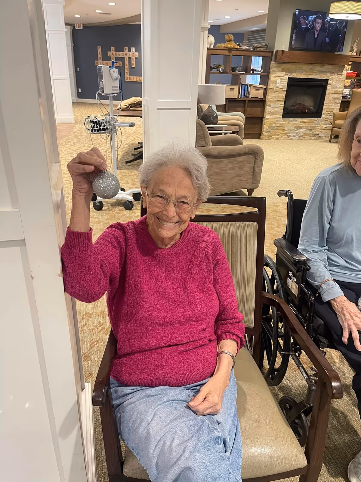 An elderly woman wearing a magenta sweater and glasses is sitting in a chair and smiling while holding up a silver Christmas ornament. Another elderly woman in a light blue top is seated next to her in a wheelchair. The background shows a cozy living room area with armchairs, a fireplace, a TV mounted on the wall, and a decorative wall with Scrabble-style letter tiles spelling words like 'LAKE ORION' and 'SMILE'.