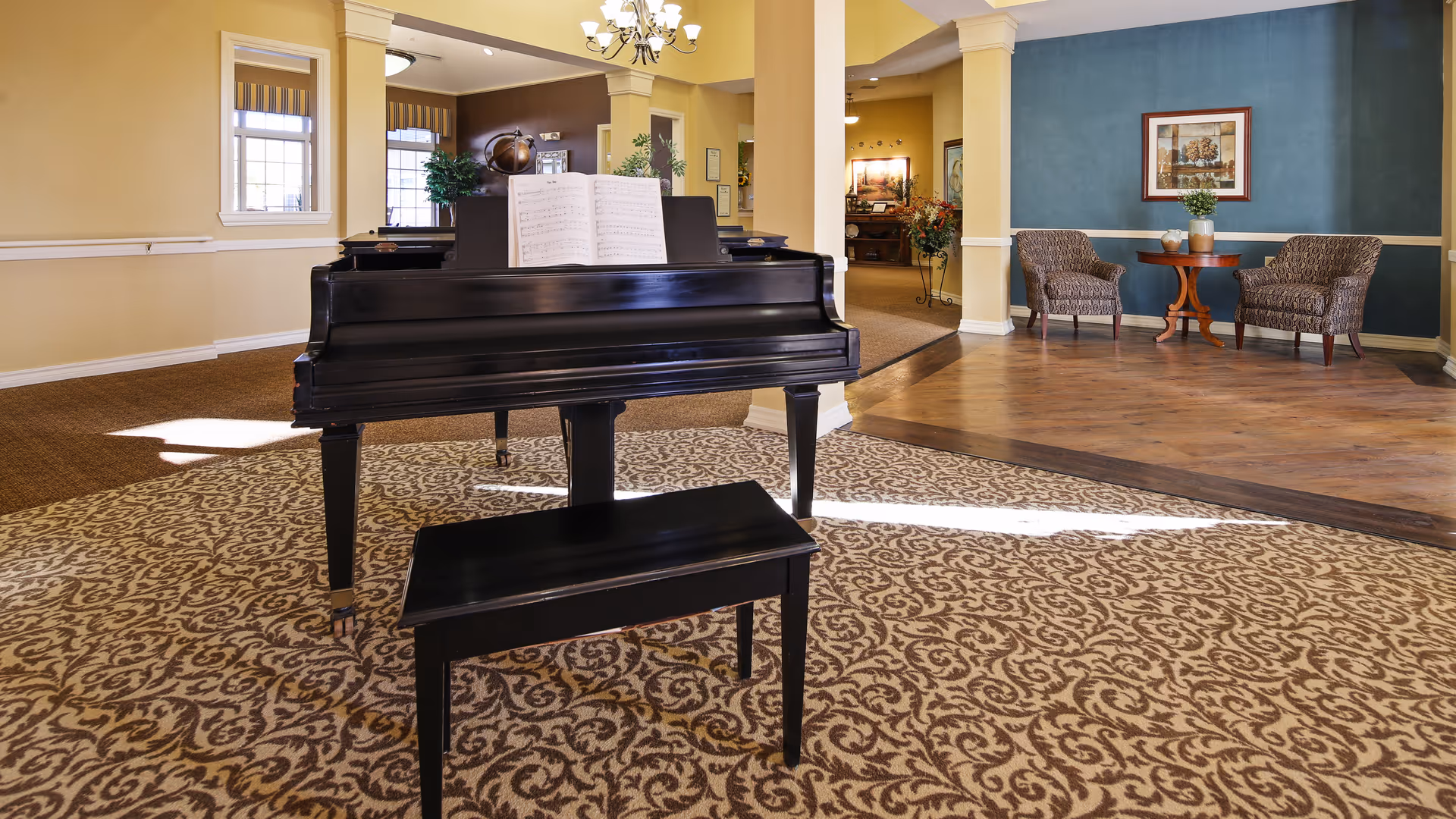 Interior view of a senior living facility common area featuring a black grand piano with an open music book on the stand, a matching piano bench, patterned carpet, and a seating area with two upholstered chairs and a small round wooden table against a blue accent wall with a framed picture above it.