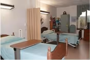 A shared bedroom in a senior living facility with two hospital-style beds, each with light blue bedding and overbed tables. A privacy curtain is partially drawn between the beds. The room has a window with green curtains, a cabinet, and some decorative items on top of the cabinet.