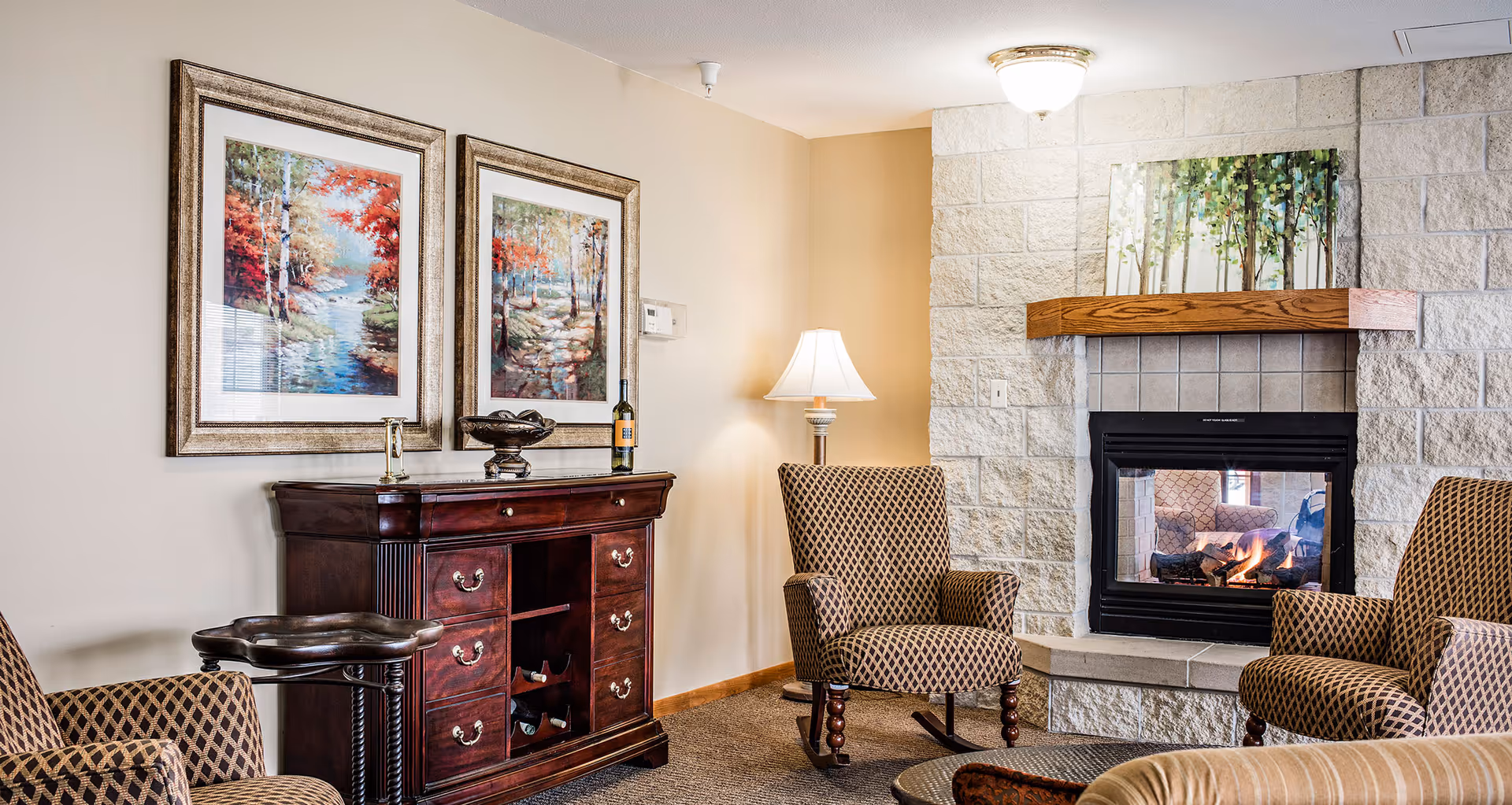 A cozy living room area with two patterned armchairs and a stone fireplace with a wooden mantle. Above the mantle is a painting of green trees. To the left, there is a dark wooden cabinet with drawers and wine storage, topped with decorative items including a bottle of wine, a bowl, and a small sculpture. Two framed paintings of autumn forest scenes hang on the wall above the cabinet. A floor lamp with a white shade stands between the cabinet and one of the armchairs.