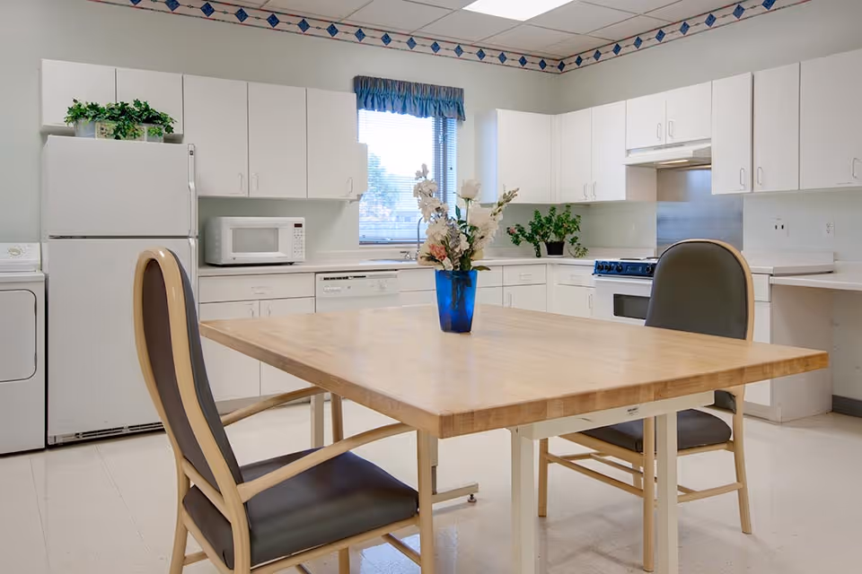 A bright kitchen area with white cabinets, a refrigerator, microwave, dishwasher, stove, and a wooden table with two chairs. A blue vase with flowers is placed on the table. There is a window with a blue valance above the sink, and some green plants on the countertop.