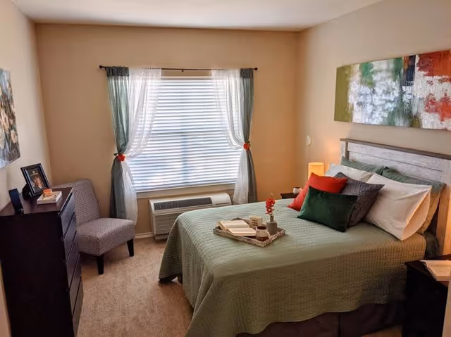 A cozy bedroom with a neatly made bed covered in a green quilt and multiple pillows in green, red, and gray. A tray with a book and a small vase with red flowers is placed on the bed. There is a window with white blinds and sheer curtains tied back with red ties. To the left of the window is a patterned chair and a dark wooden dresser with framed photos and books. A colorful abstract painting hangs above the bed, and a small lamp is on the bedside table.