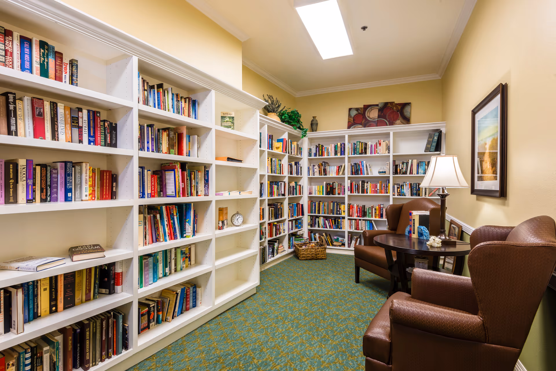 A cozy reading room with white bookshelves filled with books, two armchairs and a small table with a lamp.