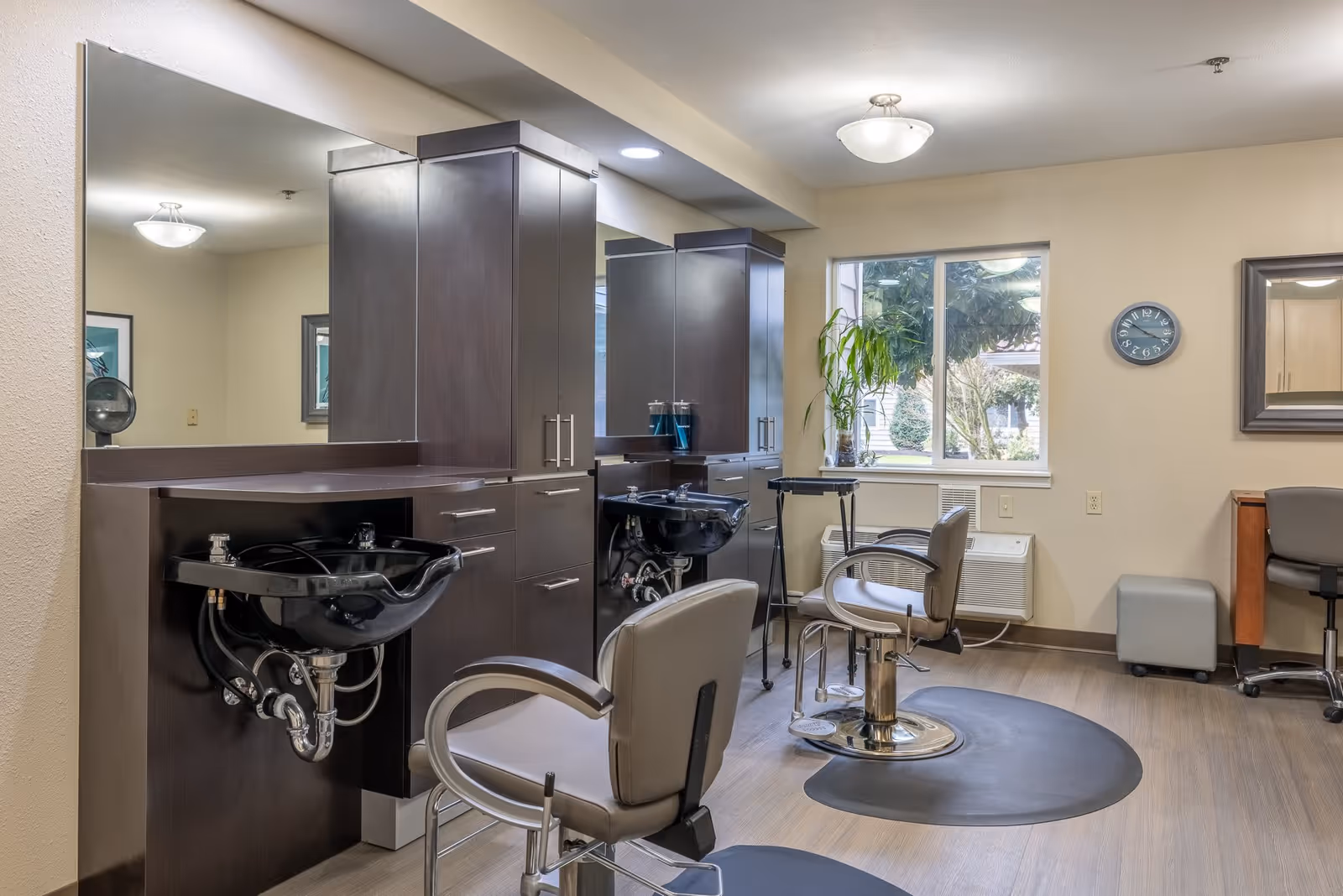Interior view of a hair salon area in a senior living facility with two salon chairs in front of black wash basins and dark wood cabinetry. There is a window showing outdoor greenery, a wall clock, a mirror, and a small desk with a chair and ottoman in the background.