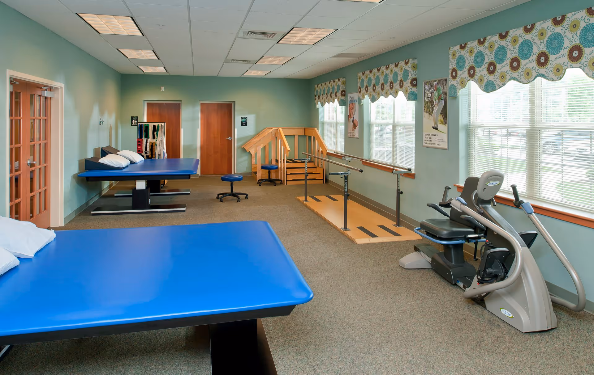 A physical therapy or rehabilitation room with two blue padded therapy tables, a stationary exercise bike, a wooden ramp with handrails, and large windows with patterned valances letting in natural light.