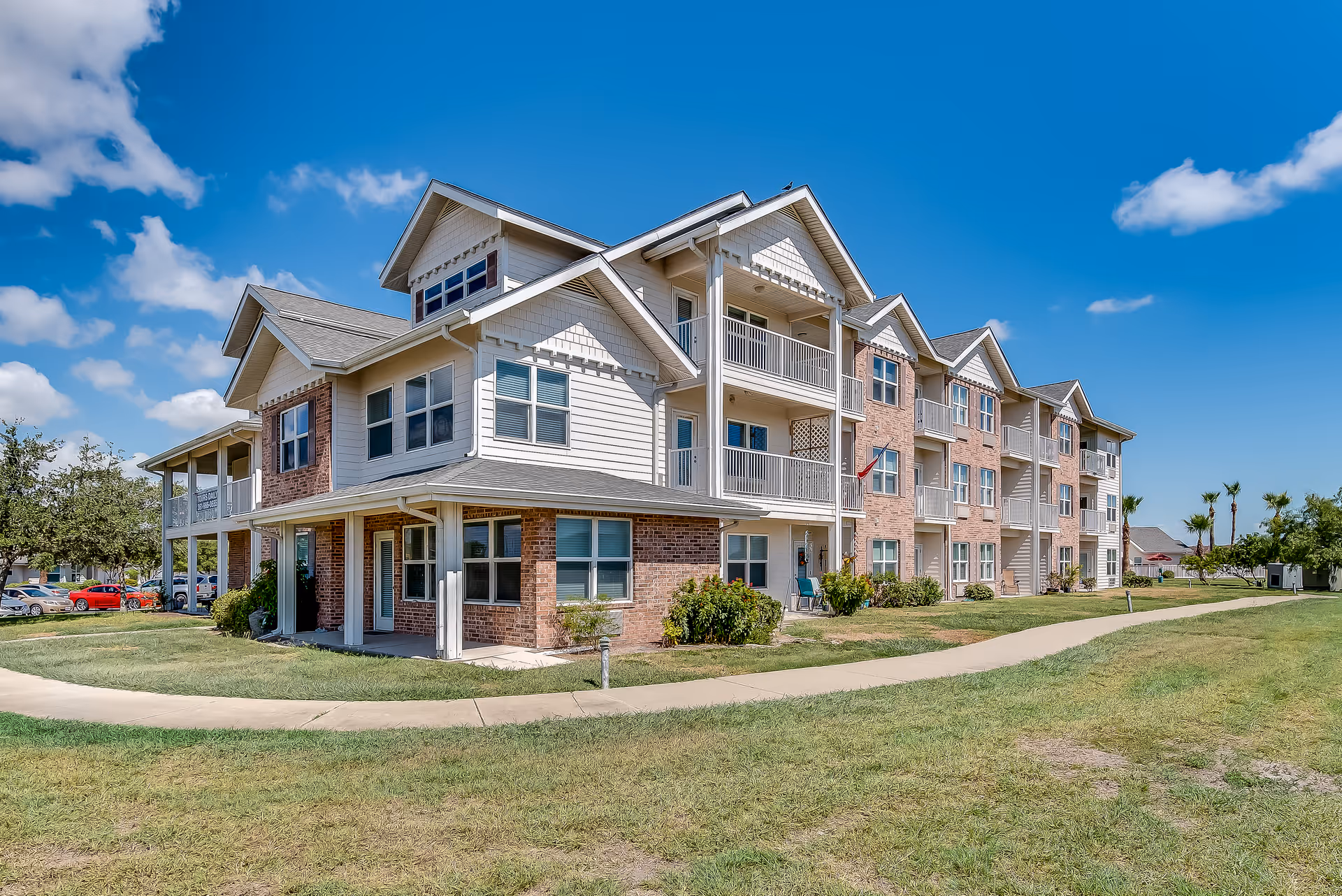 Exterior view of a multi-story senior living facility building with balconies, surrounded by a grassy lawn and a paved walkway under a blue sky with scattered clouds.