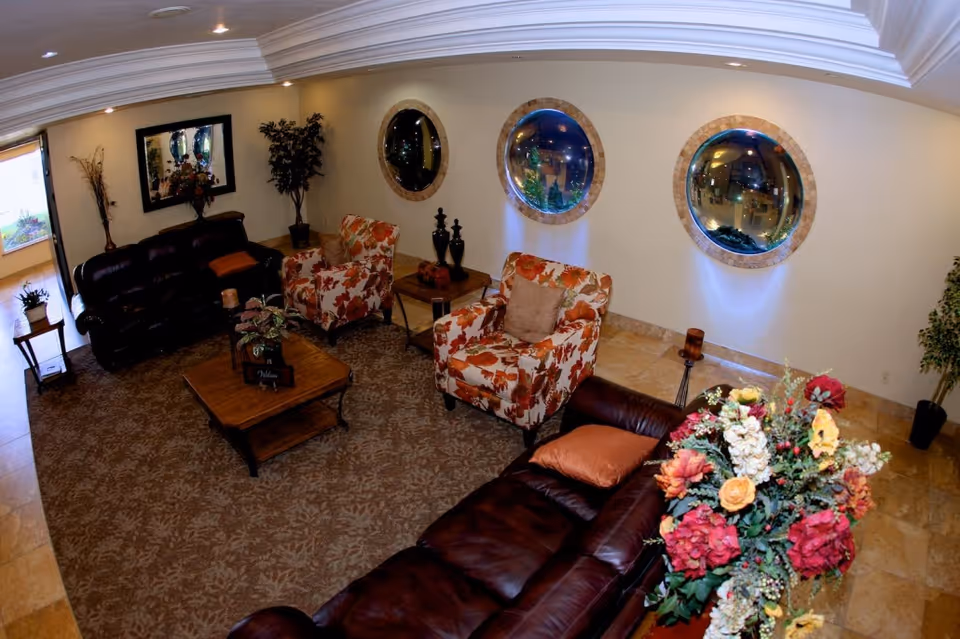 A cozy living room area with two dark brown leather sofas, two floral patterned armchairs, a wooden coffee table with a plant, and decorative plants and flowers. The walls have three round windows with stone frames and a large rectangular mirror. The floor is covered with a patterned carpet and tile.