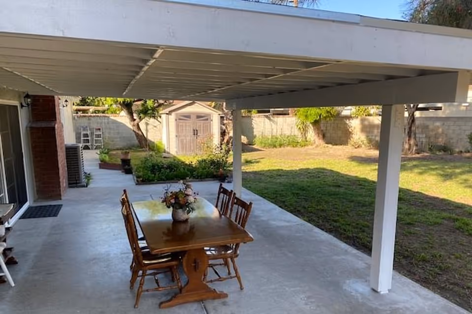 Covered outdoor patio area with a wooden table and four chairs. A flower vase is placed on the table. The patio overlooks a grassy backyard with trees, a garden bed, and a small storage shed against a concrete block wall.