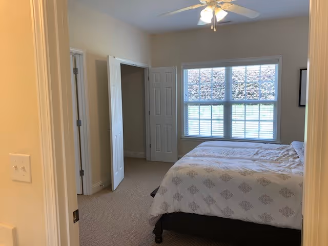 A bedroom with a bed covered in a white patterned comforter, a large window with blinds letting in natural light, a ceiling fan with lights, and an open closet with double doors.