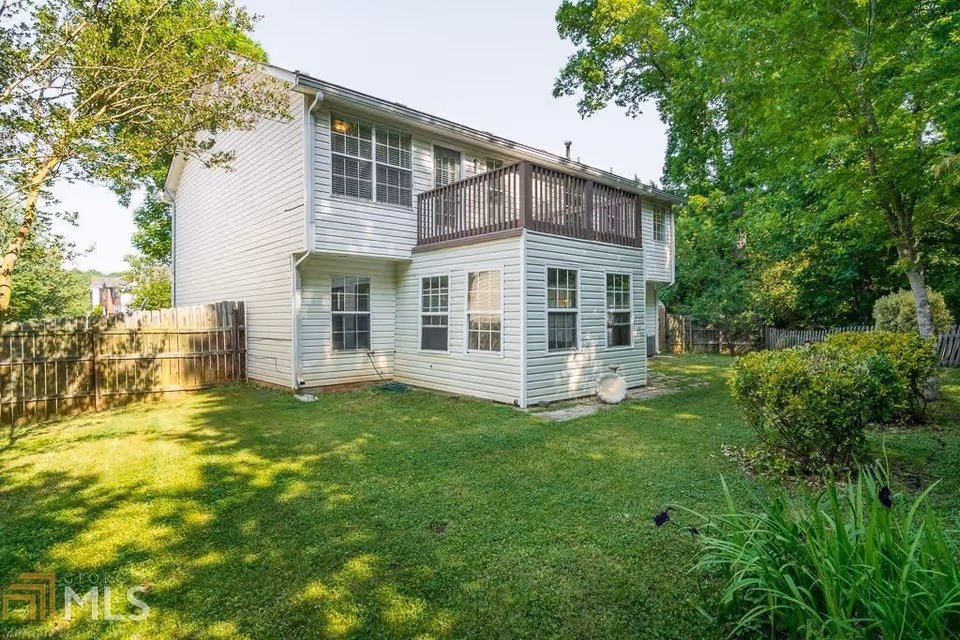 Backyard view of a two-story white house with multiple windows and a wooden balcony on the upper floor. The yard is grassy with some bushes and trees, enclosed by a wooden fence.