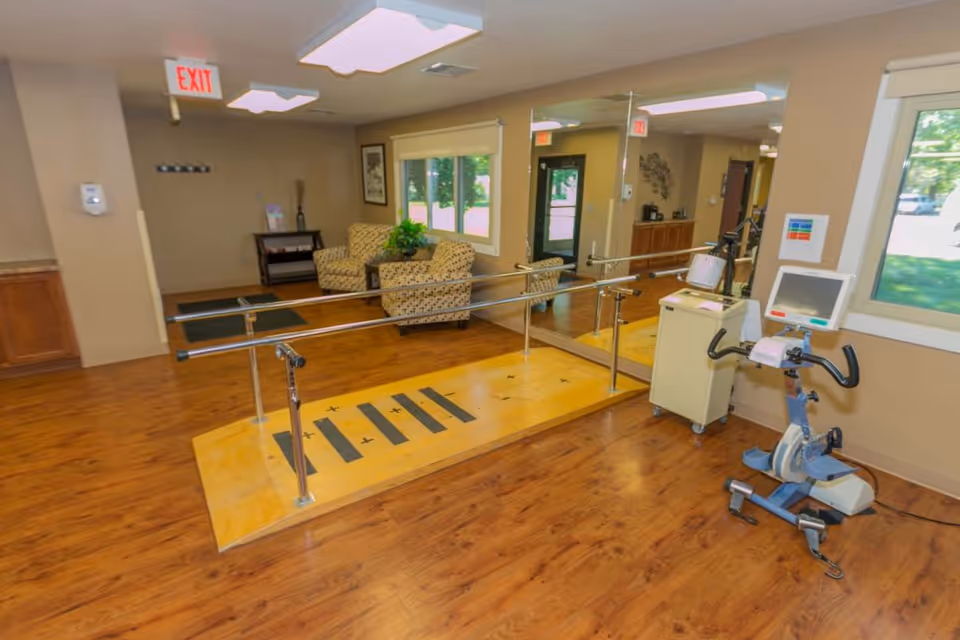 Rehabilitation/physical therapy room with parallel walking bars, an exercise bike, seating area and large mirrors.
