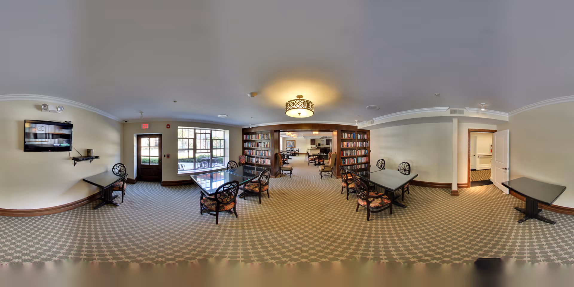 A spacious common area in Chester Street Residence featuring multiple tables with chairs arranged around them, a wall-mounted TV, large windows letting in natural light, and bookshelves filled with books. The room has patterned carpet flooring, beige walls with wooden trim, and a decorative ceiling light fixture. There is a door leading outside and an open doorway to another room.