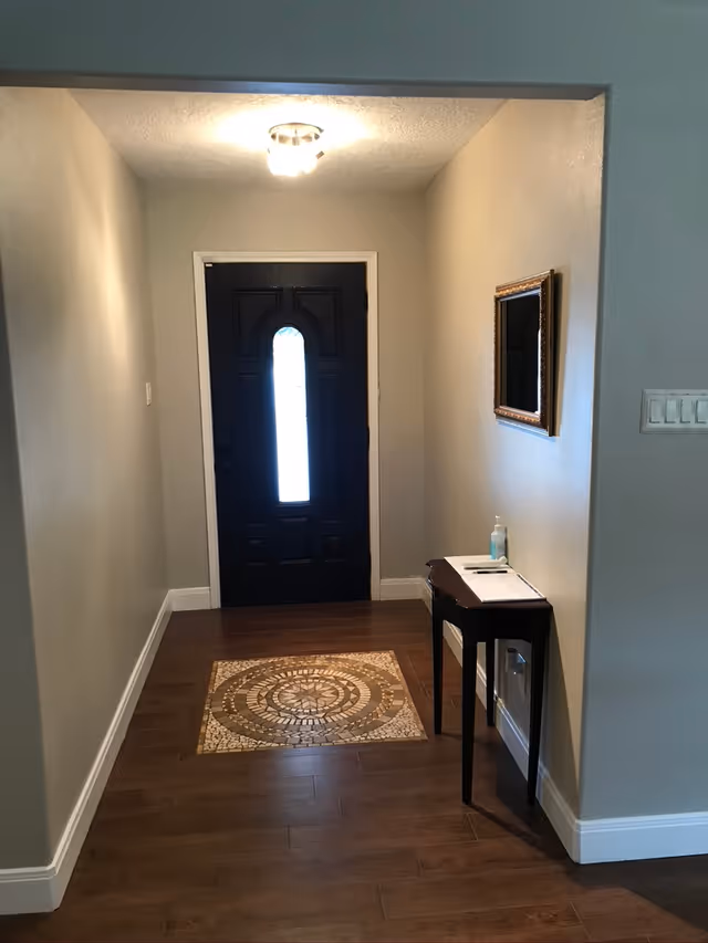 Narrow entryway with a dark front door, overhead light, a small side table with hand sanitizer, a framed mirror, and a decorative floor medallion.