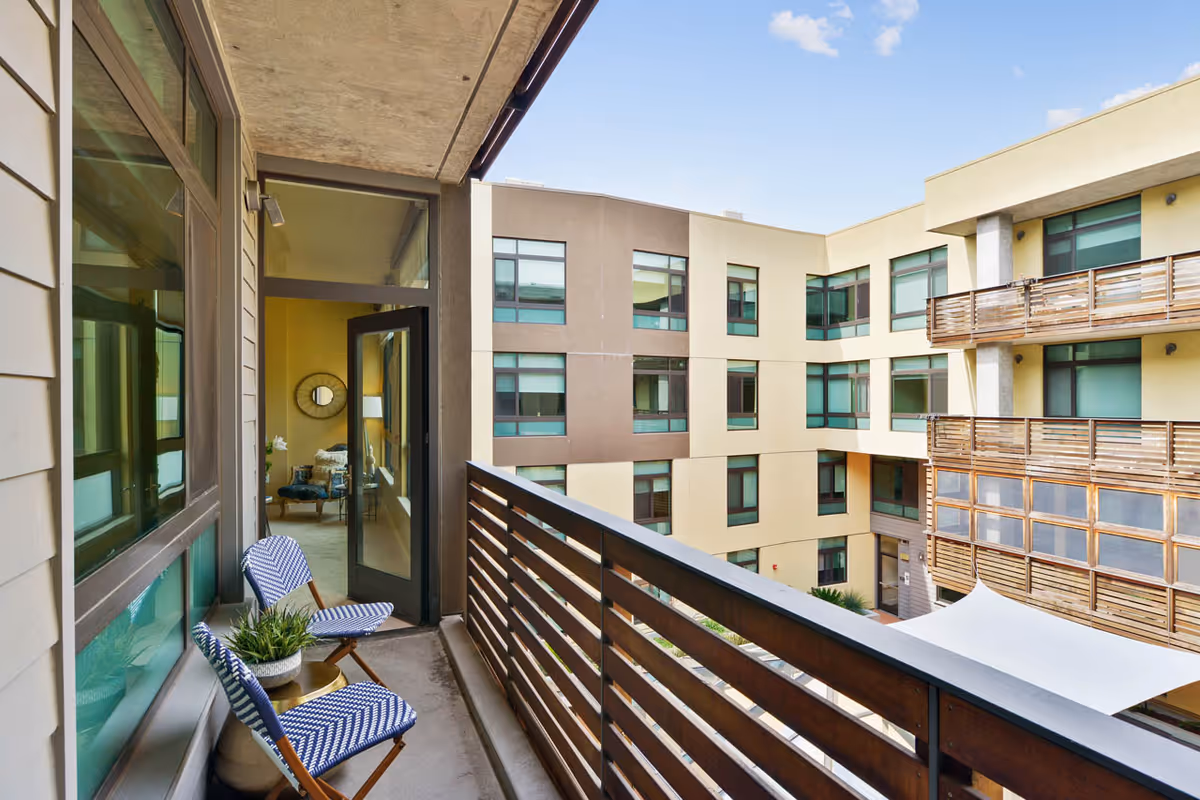 Balcony area with two blue and white patterned chairs and a small round table with a potted plant. The balcony overlooks a courtyard surrounded by a multi-story building with large windows and wooden railings. A door from the interior room opens onto the balcony, showing part of a living space with a chair and a round mirror on the wall.