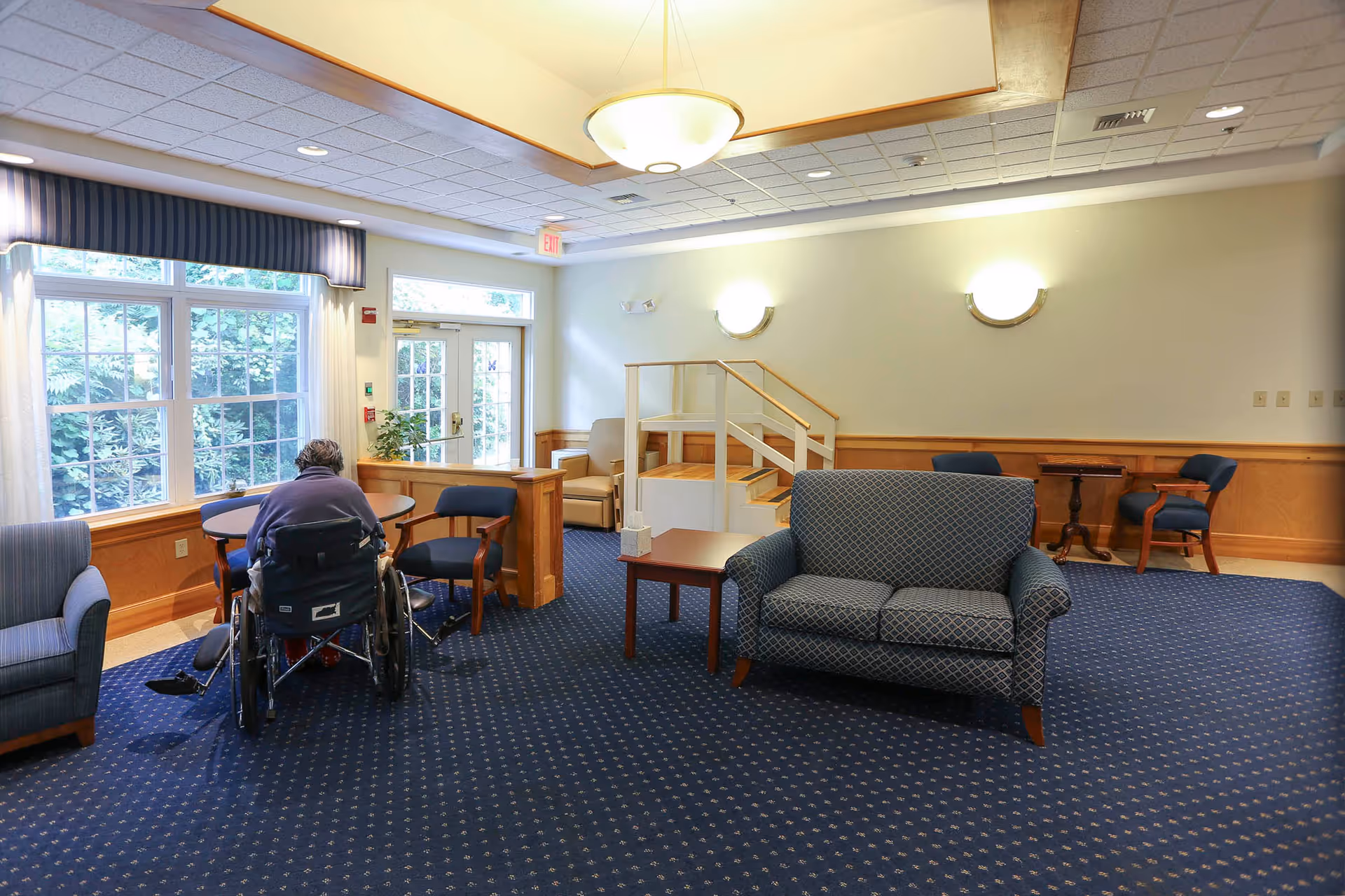Common room in a nursing facility with upholstered sofas and chairs, a person in a wheelchair seated at a table, and large windows.