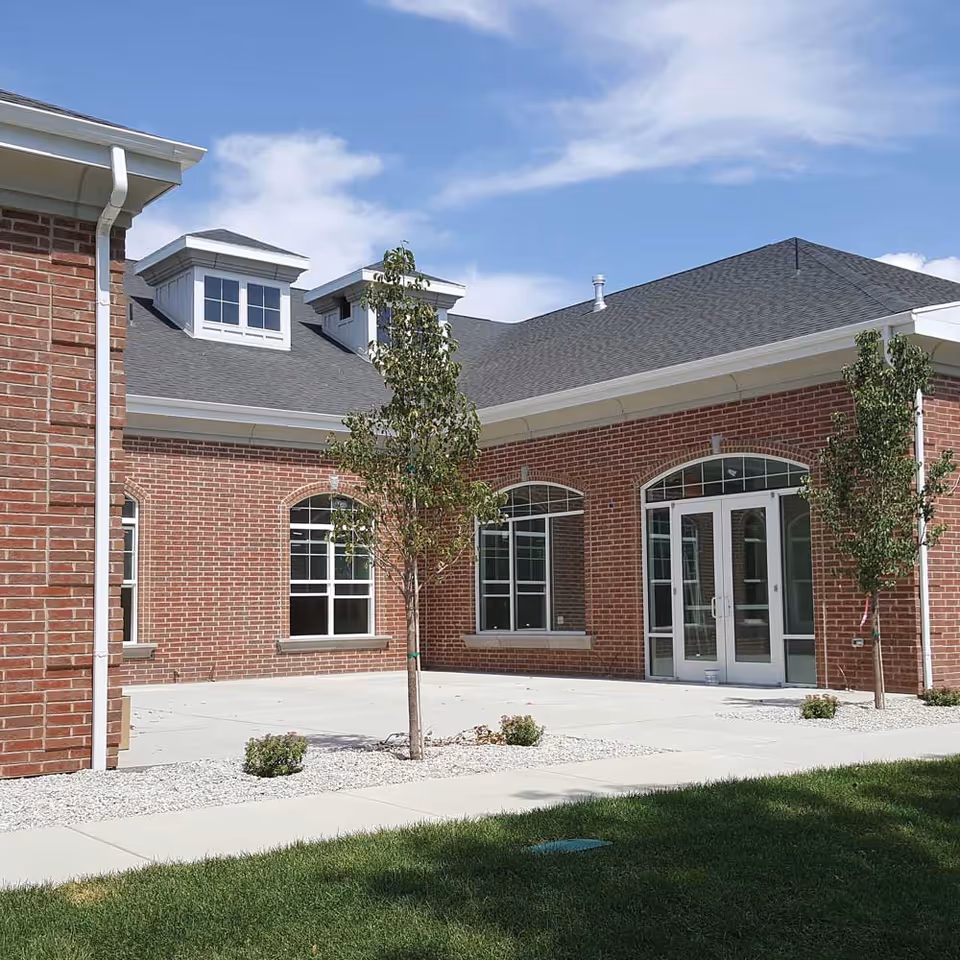 Exterior view of a brick building with large windows and double glass doors, surrounded by a concrete patio, small trees, and green grass under a partly cloudy sky.