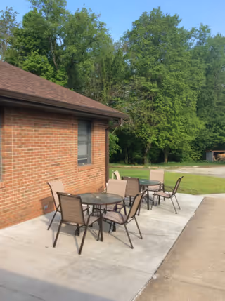Outdoor patio area next to a brick building with three round glass tables and several brown chairs arranged around them. The patio is on a concrete surface and surrounded by green grass and tall trees in the background under a clear blue sky.