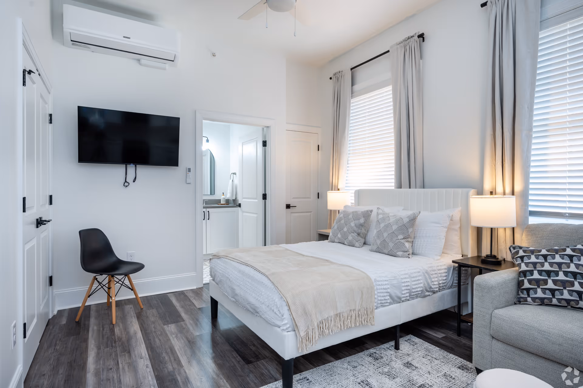 A modern bedroom with a white bed featuring multiple pillows and a beige throw blanket. There are two windows with white blinds and light gray curtains. A gray armchair with a patterned pillow is next to the bed, along with a black side table and lamp. A black chair is placed against the wall beneath a mounted flat-screen TV. The room has dark wood flooring and white walls. An open door reveals a bathroom with a vanity and mirror.