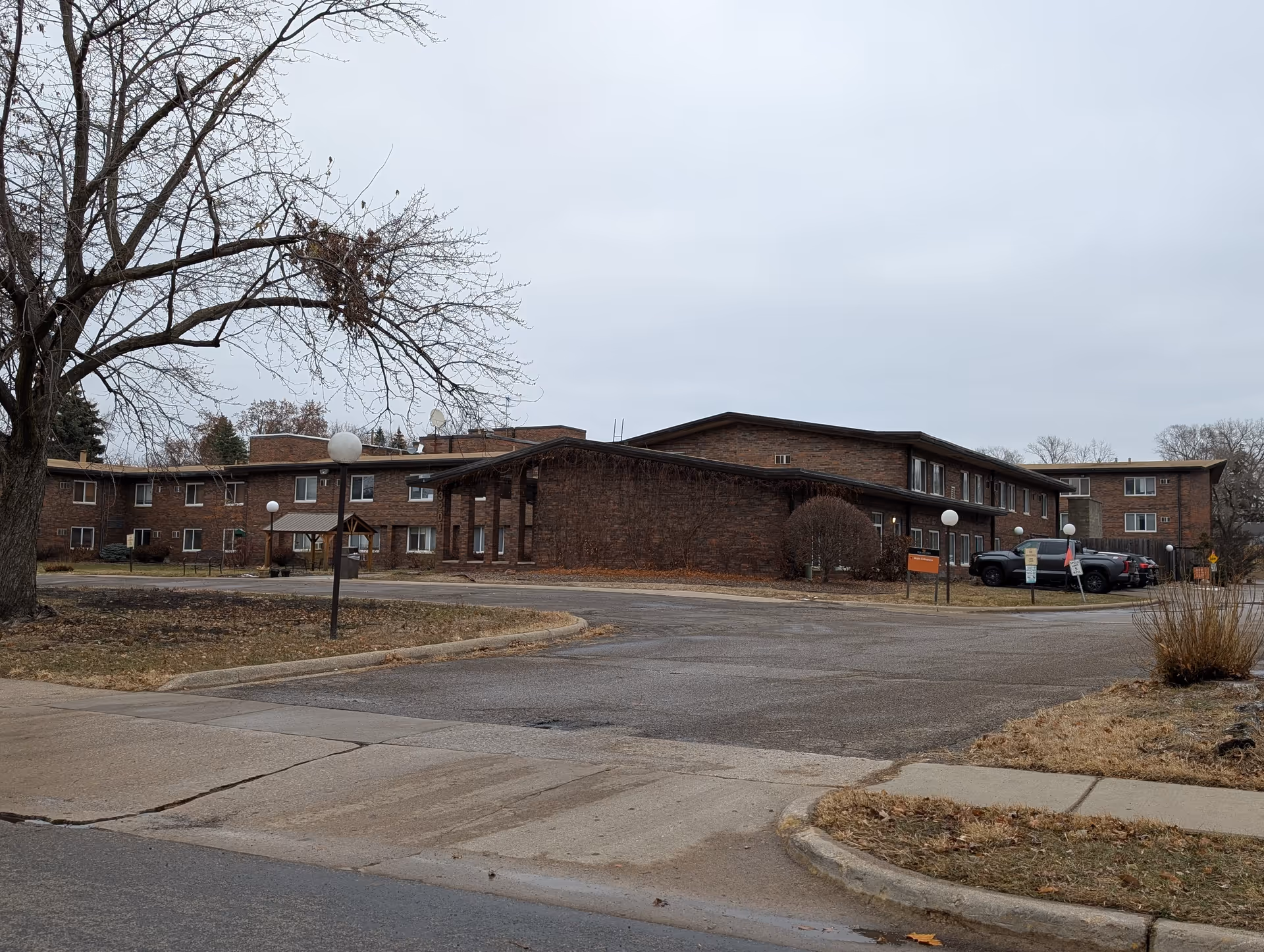 Two-story brick senior living building with a parking lot, bare trees, and overcast sky.