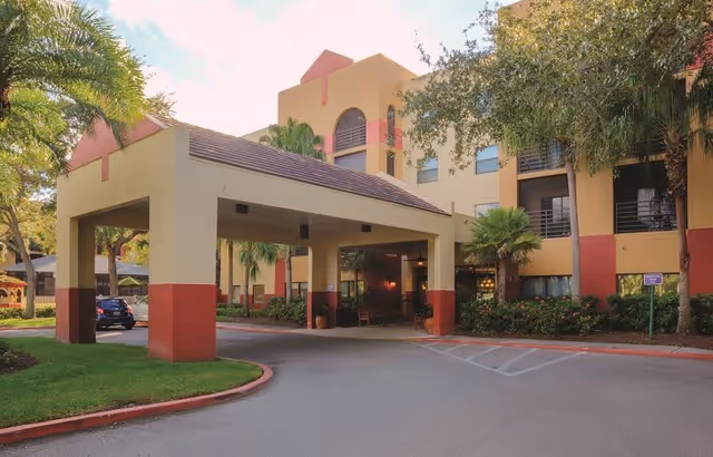 Exterior view of a multi-story assisted living facility with a covered entrance driveway, surrounded by palm trees and landscaping under a partly cloudy sky.