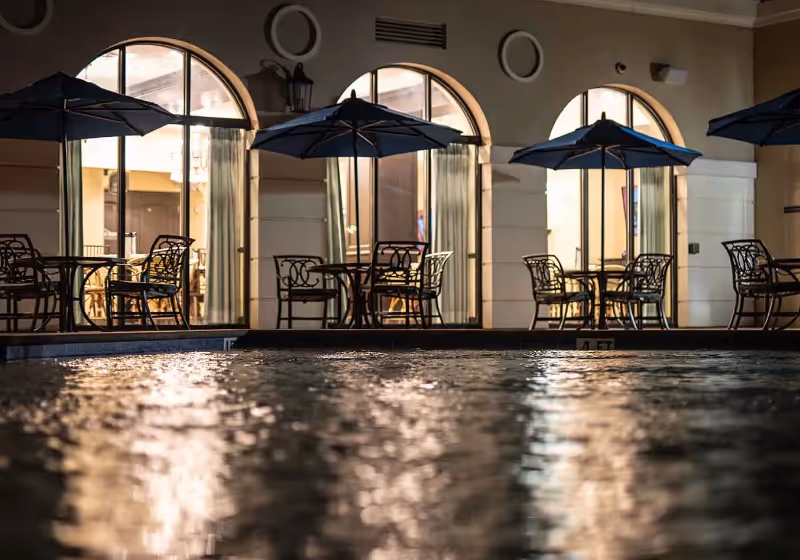 Night view of a pool with reflections in the water and a patio lined with tables, chairs, umbrellas, and arched windows.