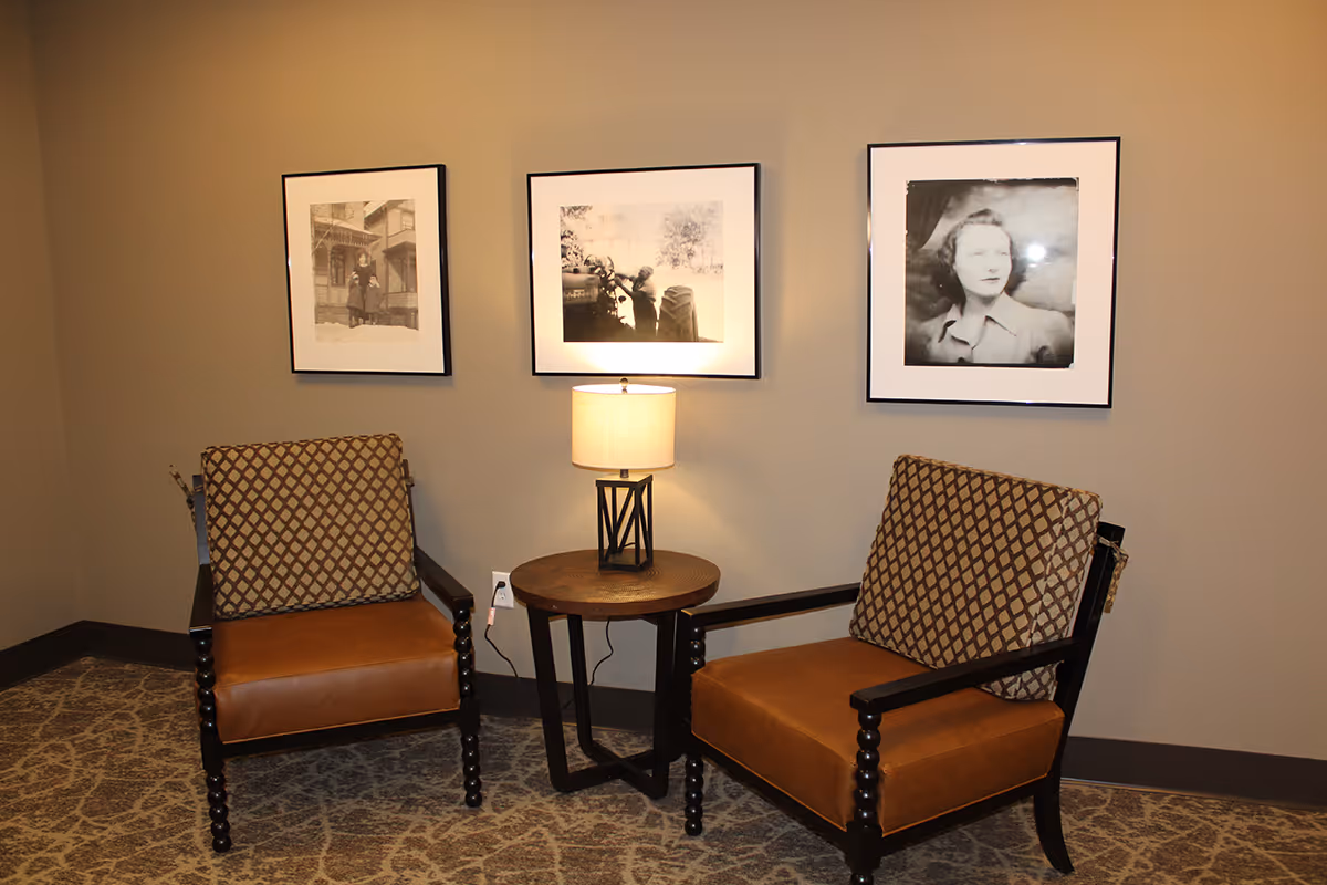 Two brown cushioned armchairs with patterned back cushions are placed on either side of a small round wooden table with a lamp on it. Behind the chairs, three black and white framed photographs hang on a beige wall. The floor is carpeted with a patterned design.