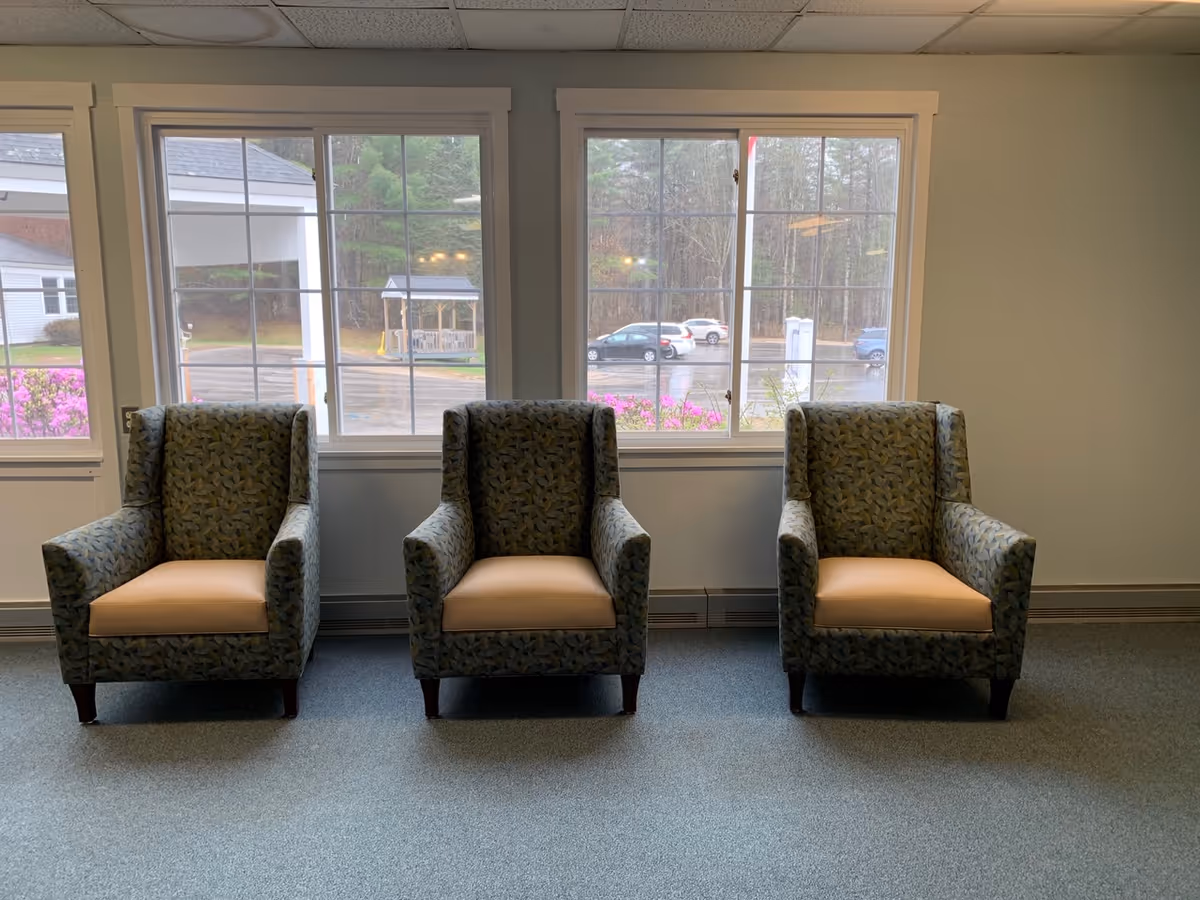 Three patterned armchairs with beige cushions arranged in a row in front of three large windows showing a view of a parking lot and trees outside.