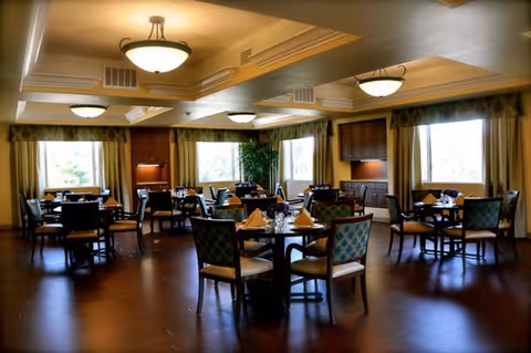 Well-lit dining room with multiple round tables set with napkins and chairs, large windows and decorative ceiling lights.