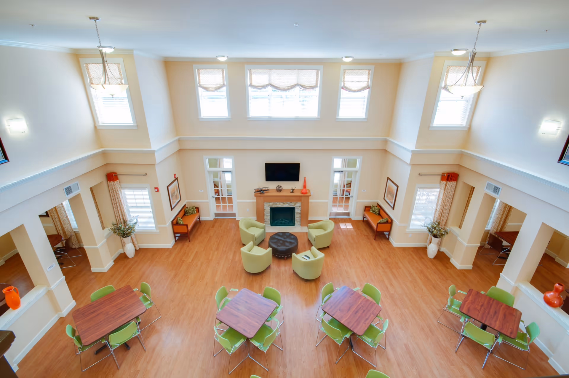 A spacious, well-lit common area with high ceilings and large windows. The room features four square wooden tables each surrounded by green chairs, a seating area with four green armchairs around a round black ottoman, and a fireplace with a mounted TV above it. The walls are light beige, and there are decorative vases and framed pictures on the walls. The floor is wood, and there are two benches with cushions along the far wall.