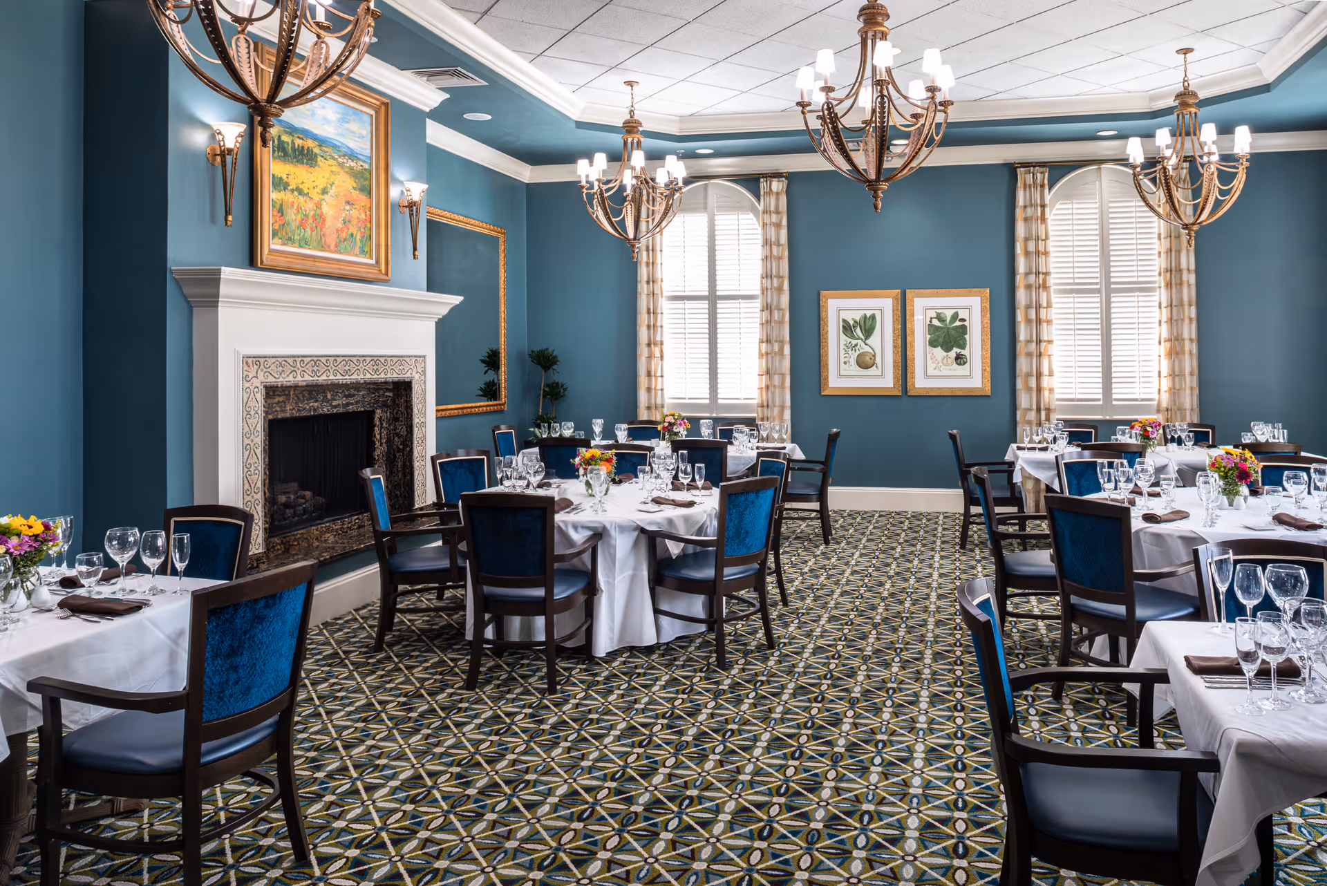 Elegant dining room with round tables set with white linens and glassware, blue walls, chandeliers, patterned carpet and a fireplace.