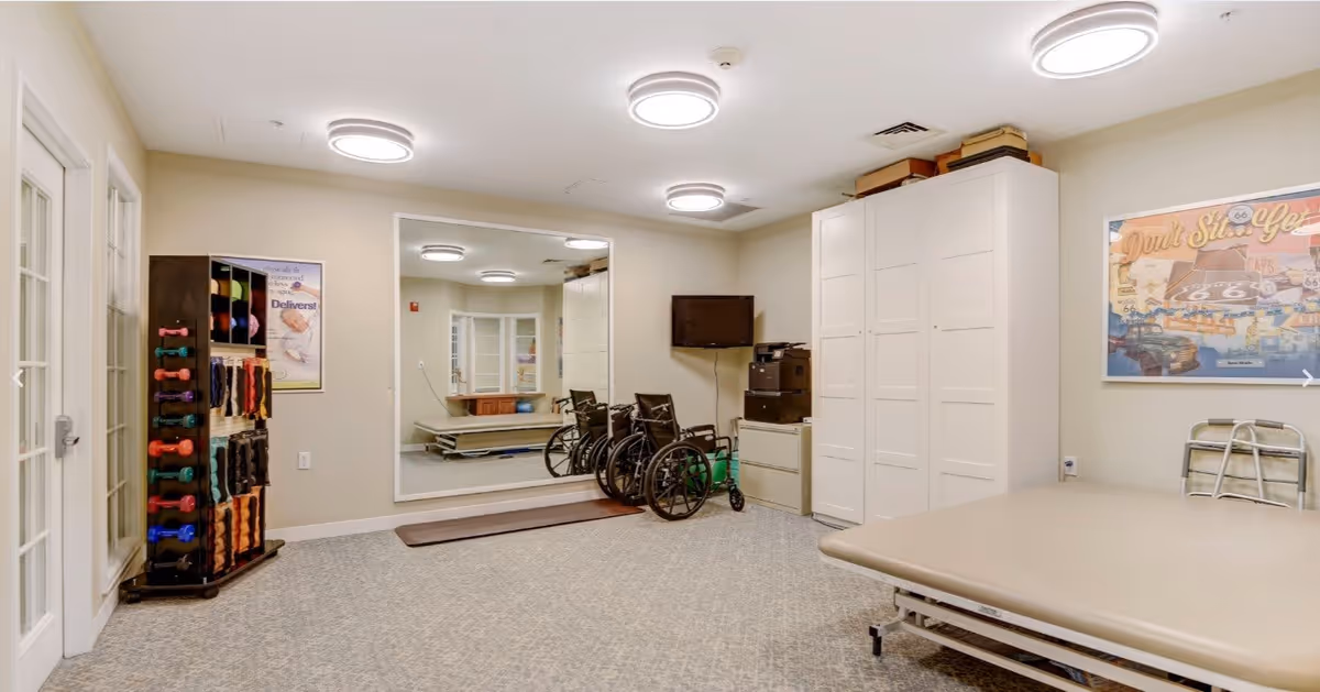 A therapy or rehabilitation room in a senior living facility with a large mirror on the wall, several wheelchairs, a therapy table, a rack of colorful hand weights, and a walker. The room has beige walls, carpeted floor, and modern circular ceiling lights.