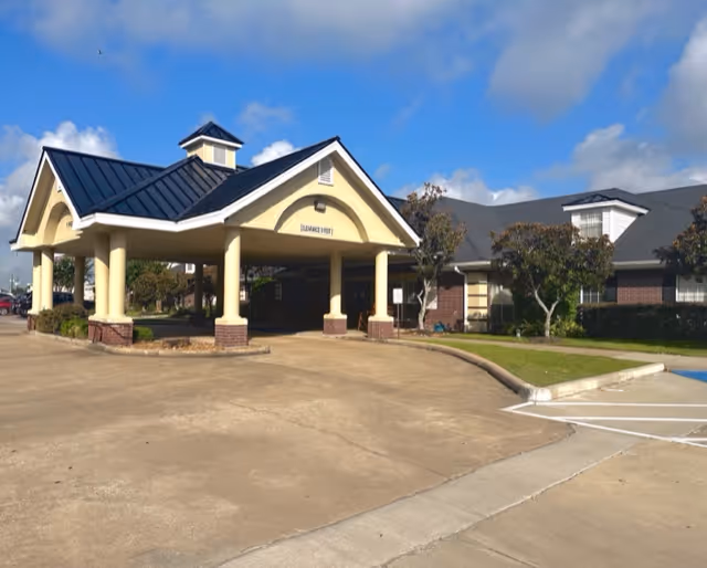Exterior view of Park Manor of South Belt showing the front entrance with a covered drop-off area supported by columns, a paved driveway, and surrounding greenery under a partly cloudy sky.