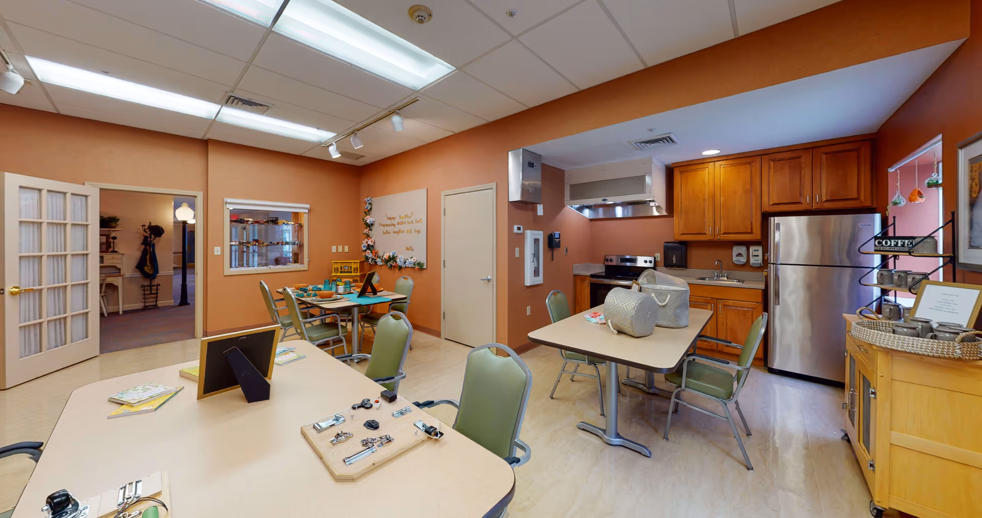 Interior view of a communal kitchen and activity area in a senior living facility. The room features beige walls and ceiling lights. There are tables with chairs around them, some tables have activity boards and baskets on top. The kitchen area includes wooden cabinets, a stainless steel refrigerator, a stove with a hood, and a sink. A small coffee station is set up on a wooden cart near a window with hanging decorative items.