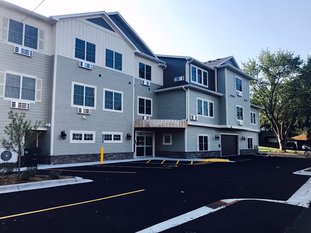 Exterior view of a modern three-story senior living facility building with multiple windows, air conditioning units, and a parking area in front. There are trees and landscaping around the building under a clear sky.